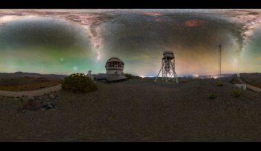 A glowing panorama shows jagged shapes of blue and purple of the Milky Way in a green and yellow night sky. Below are metal domed buildings with a barren landscape around them