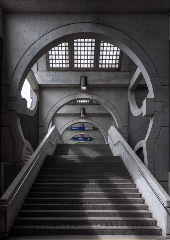 A wide concrete staircase leads upward, flanked by white railings. Overhead, geometric arches, circular cutouts, and a grid-patterned skylight create a modern architectural look. Light and shadows play on the steps.