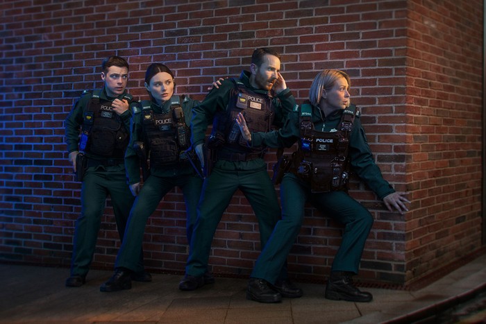 4 police officers in uniform pressed up alongside a brick wall all looking around the corner.