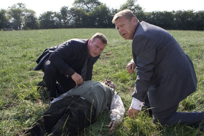 Dalziel and Pascoe lean over and inspect an unconscious man lying in a grass field.