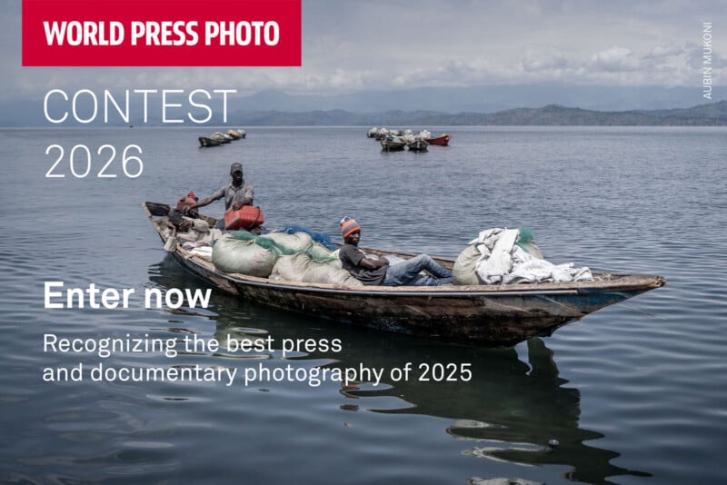 Two men sit in a wooden boat filled with sacks on a calm lake, with distant hills and another boat in the background. Text promotes the 2026 World Press Photo Contest for press and documentary photography.