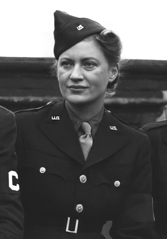 A black and white photograph of a woman in a military uniform. She is wearing a cap and a tie, with buttons and insignia on her jacket. The background is blurred, focusing attention on the subject's face and attire.