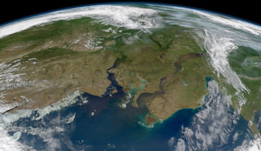A satellite view of a portion of Earth shows the planet’s curved horizon at the top of the image. Green and brown landmasses are cut by two large, blue, winding rivers that empty into the ocean in the foreground. Clouds are visible on the edges of the image.