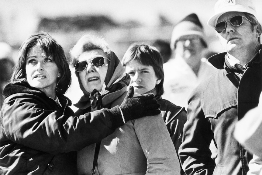 Black-and-white photo of four people looking up. One has an arm around another and their faces are apprehensive.