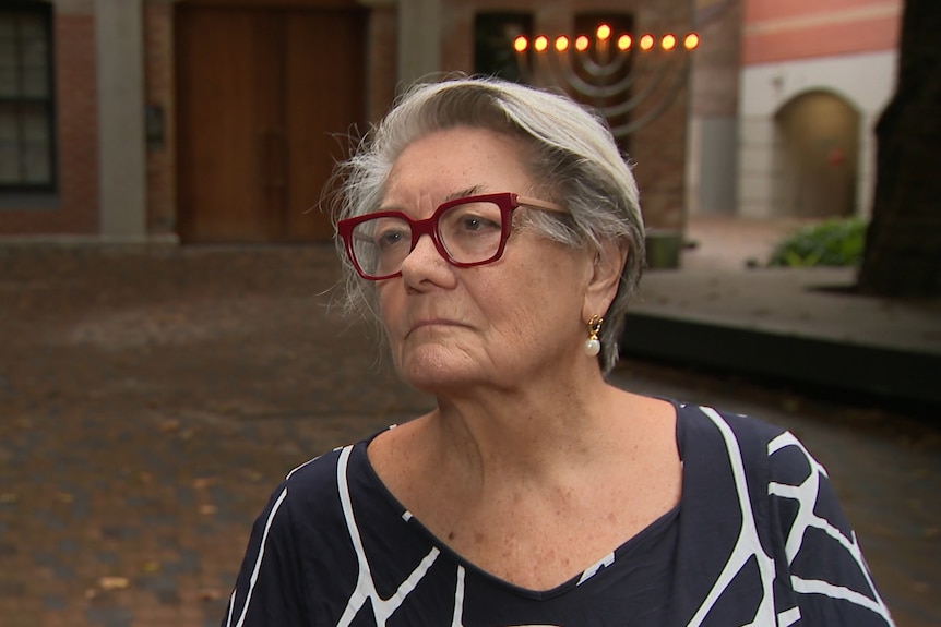 Ms Betts wearing maroon rimmed glasses wither hair up standing in front of a brick building and a menorah.
