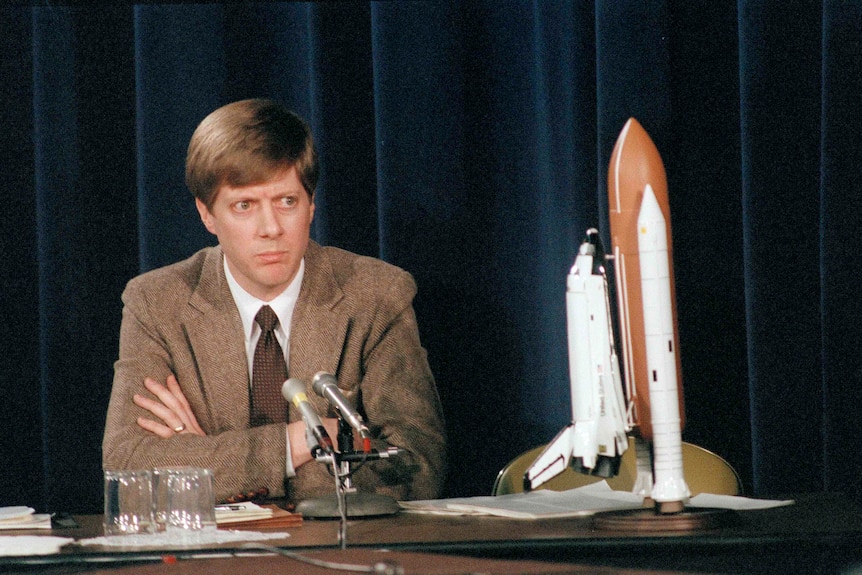 A concerned young man of about 30 in jacket and tie at a desk behind a microphone, a space shuttle model beside him.