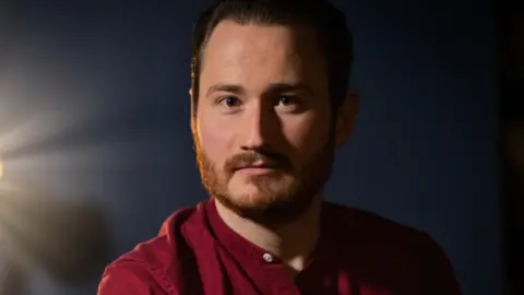 A man wearing a red buttonless shirt, lit from the side by a bright, warm light, standing against a dark blue background