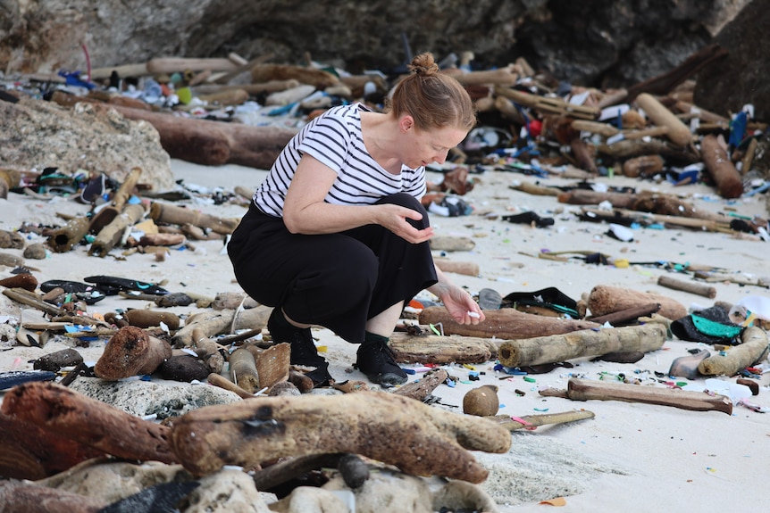 A woman wearing a striped shirt and pants crouches on a beach and picks up a white piece of plastic among many pieces of debris.
