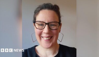 Lucy is looking at the camera and can be seen from the shoulders up. She's wearing large square-hooped earrings and smiling, with her hair back.