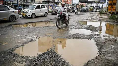 Getty Images In this photograph taken on October 22, 2025, commuters ride along a street riddled with potholes in Bengaluru. Home to nearly 12 million people and state capital of Karnataka, Bengaluru is the "Silicon Valley" of the world's fifth biggest economy -- hosting thousands of start-ups, outsourcing firms, and global tech giants from Google to Microsoft. Yet its flagship Outer Ring Road (ORR) business district is clogged with traffic, pocked with potholes, and often flooded during the monsoon.