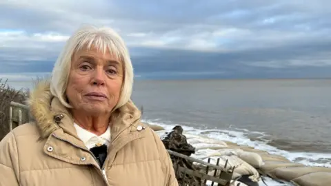 Richard Daniel/BBC A head and shoulders image of Maureen Jones standing on the Thorpeness coastline against a backdrop of the sea and sea defences.