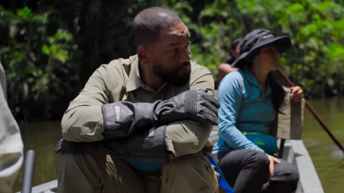 actor will smith in khakis seated with arms folded on is knees in a canoe rowing down a tropical river