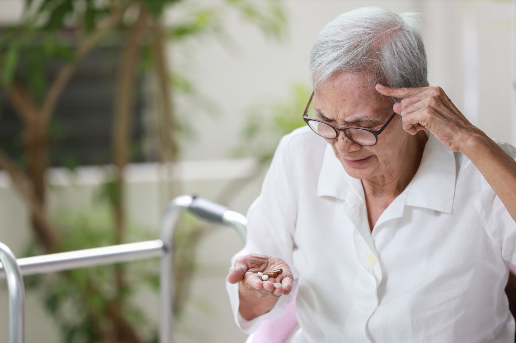 An elderly person with gray hair and glasses looks confusedly at pills in her palm, with one hand touching her temple.