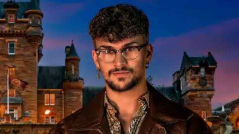 Cody Burridge/BBC/Studio Lambert Jack is in the middle looking at the camera. He has short brown curly hair and is wearing glasses, a patterned shirt and a brown leather jacket. He is stood in front of a castle. 