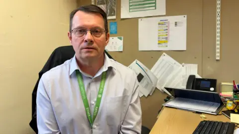 Des Gorman pictured in a white button down short and a green lanyard. He has short dark hair and glasses.