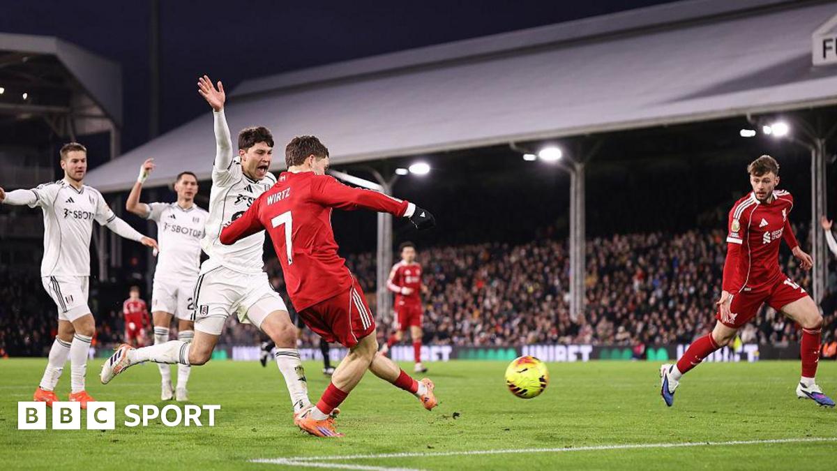 Liverpool's Florian Wirtz scores against Fulham. He is in the penalty area and has struck a yellow ball with his left foot. Liverpool's kit is red and he is wearing orange boots. A Fulham player, wearing all white, is next to him and is raising his right arm to call for offside. Several other players can be seen in the background along with thousands of fans in one of the stands.