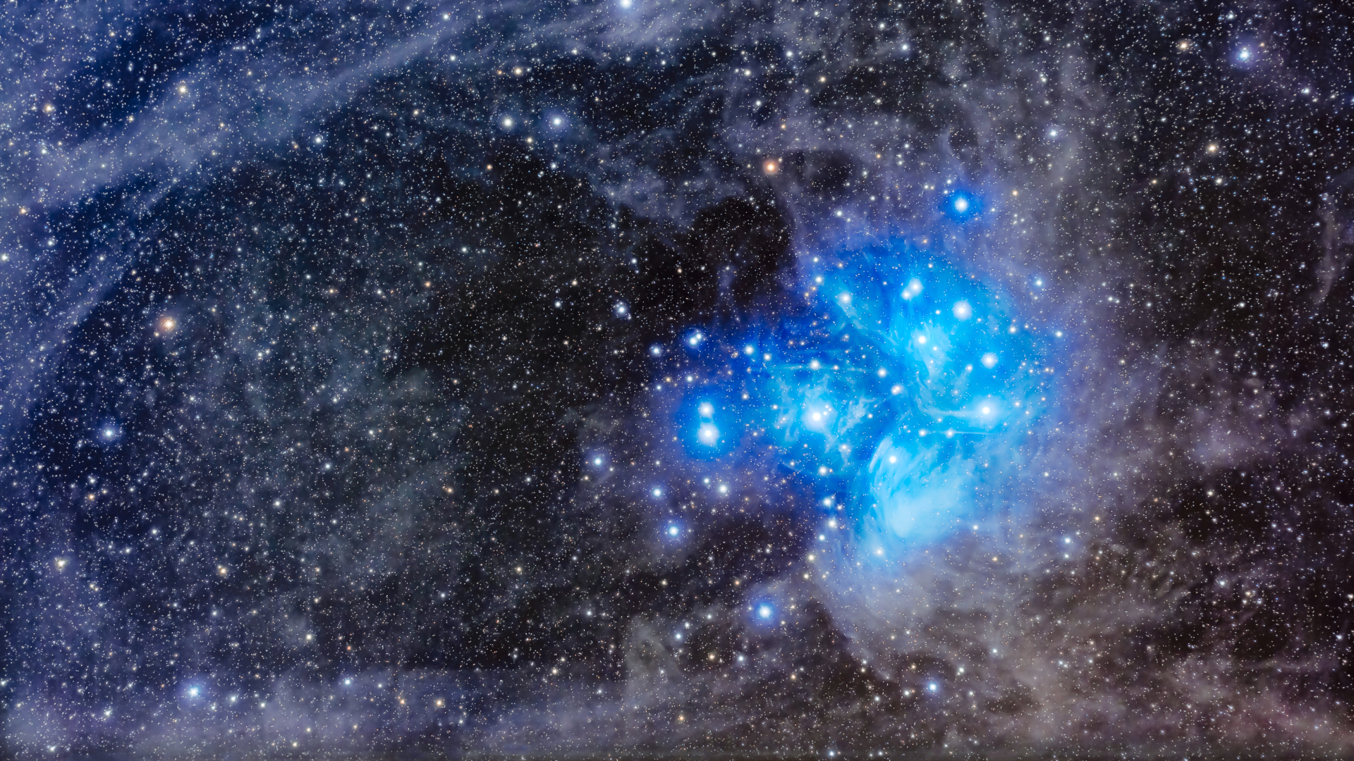 A collection of blue-white stars are pictured surrounded by a diffuse blue glow against the dimmer starfield beyond, which is threaded with wispy clouds.