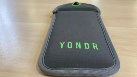 A grey pocket sleeve wallet sits on a wooden table. On it is the company's name Yondr in green colouring. It is designed to store a mobile phone device with a magnetic seal which can be reopened at a later time or date