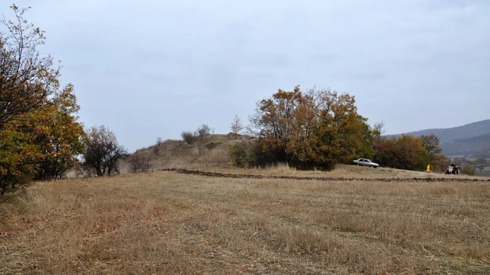  A small grass-covered mound with dotted trees against a landscape. 