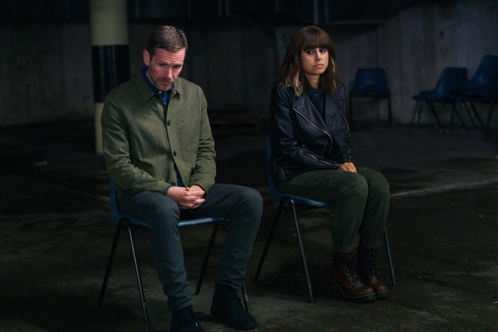 Shaun Evans as John Hughes and Zahra Ahmadi as Mehreen Askari-Evans in Betrayal, sat together on hard chairs in a darkened room.