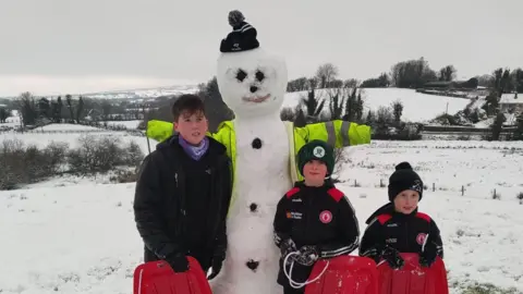 BBC WEATHER WATCHER - Gortin Glen Glamping Three young boys standing next to a snowman they made. They are all wrapped up in gloves, hats and coats, and are holding onto red sleighs.
