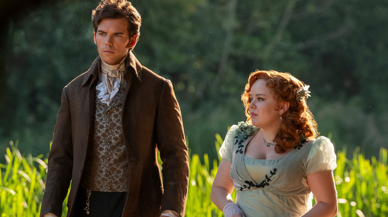 Luke Newton as Colin and Nicola Coughlan as Penelope dressed in period costumes walking outside in Bridgerton
