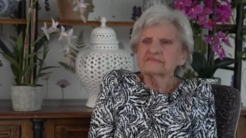 BBC A woman with grey wears a black and white top and sits in front of an oak dresser with an orchid and vase on it.