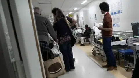 Patients lie on trolleys along a hospital corridor while people stand and walk between them.