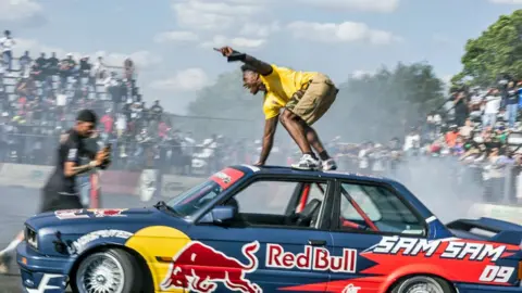 AFP/Getty Images YouTuber and online streamer Darren Jason Watkins Jr, better known as IShowSpeed, holds on to the top of the car of Samkeliso Samsam Thubane's, also known as Sam Sam and an official Red Bull athlete, in the south of Johannesburg, on 2 January 2026