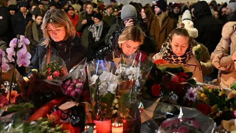 Getty Images People gather at the scene of a deadly bar fire in Switzerland. They are stood in front of a huge pile of flowers and candles.