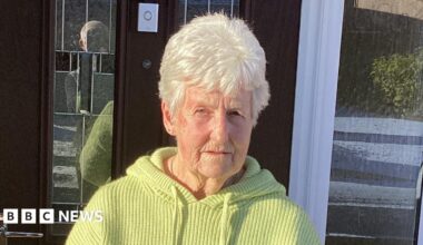 A woman with white hair and wearing a yellow top standing outside a front door. It is a sunny day and there are shadows in the background.