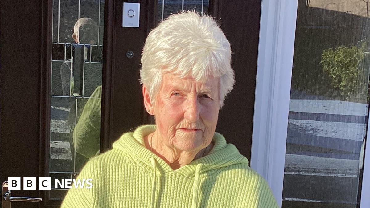 A woman with white hair and wearing a yellow top standing outside a front door. It is a sunny day and there are shadows in the background.