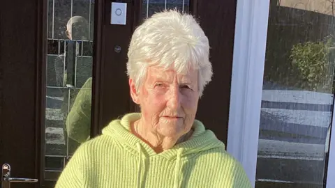 Tony Brown A woman with white hair and wearing a yellow top standing outside a front door. It is a sunny day and there are shadows in the background.