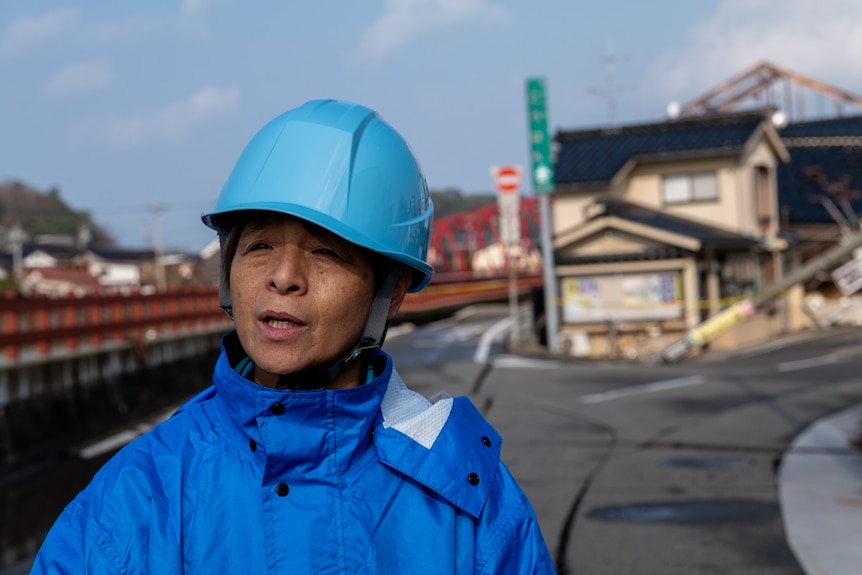 A woman in a helmet and jacket stands near a small building next to a bridge.
