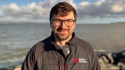 A man standing in front of Belfast Lough. He has short brown hair and a short brown beard. He is wearing black rimmed glasses and a black jacket with Belfast Harbour written on it in white.