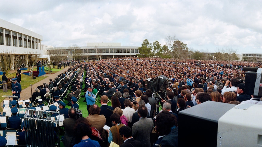 Wide-format photo showing a huge crowd seated in rows on a lawn, with a military band and TV cameras in foreground.