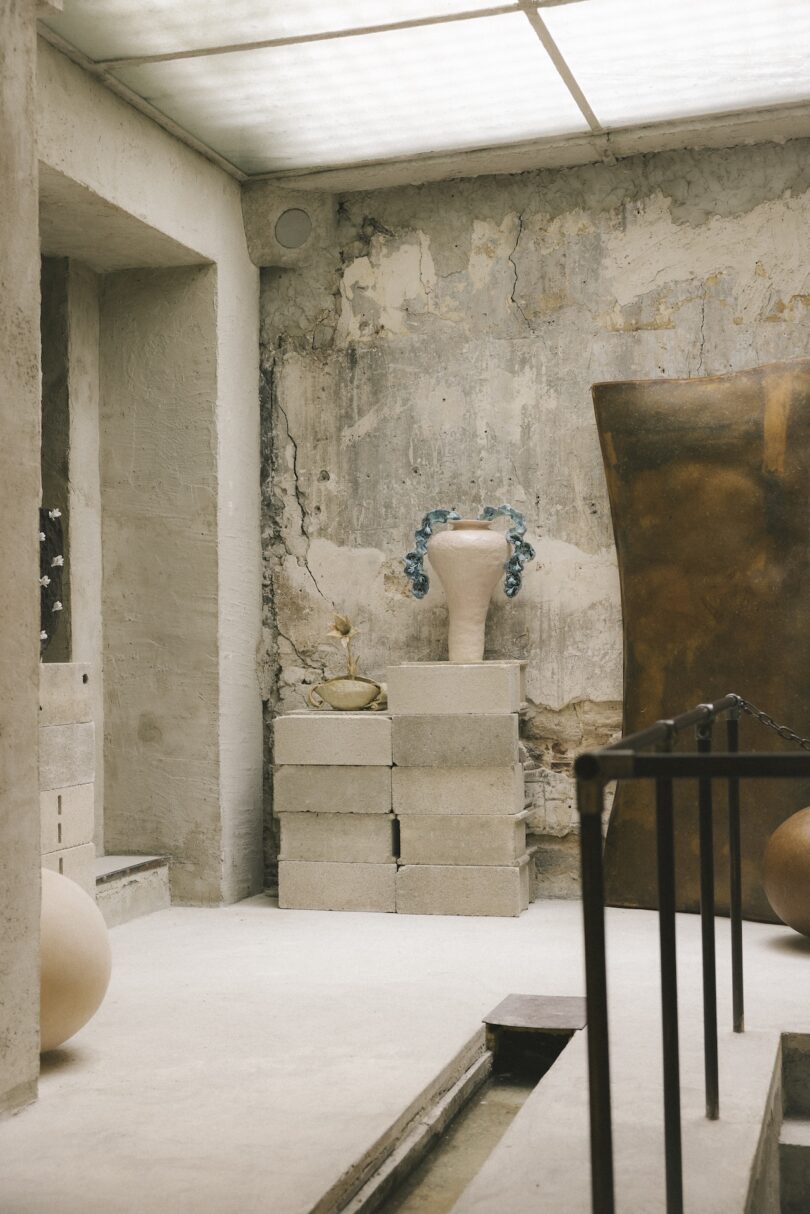 A large ceramic vase with blue decorations sits on stacked concrete blocks in a minimalist, unfinished room with rough walls and art objects.