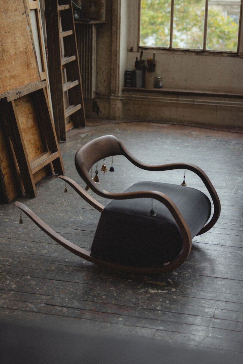 A modern wooden rocking chair with a black cushion sits on a worn wooden floor in an artist's studio, near a window and art supplies.