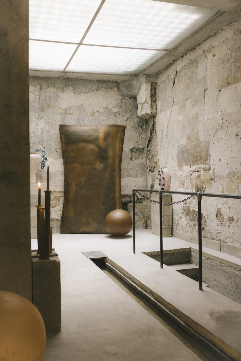 A minimalist room with rough, textured walls, abstract sculptures, a bronze sheet, candles, and a sunken rectangular trench in the floor.