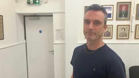 Amy Holmes/BBC A man wearing a navy T‑shirt stands inside a hallway decorated with framed portraits on the wall. To the left is a white door beneath a green emergency exit sign, and a few chairs are positioned along the wall on the right.
