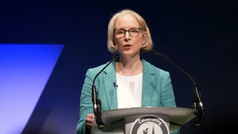 PA Media Reynolds stands at a podium, addressing the conference, in a green jacket and lit up in front of a purple background
