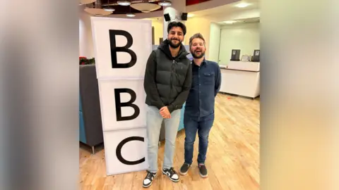 Ekam Dhaliwal A young man with black hair and facial hair is wearing a black top, black gilet and blue jeans. He is standing next to a shorter man with dark brown and grey hair and facial air, and a blue top and jeans. Next to them are three blocks that spell out BBC