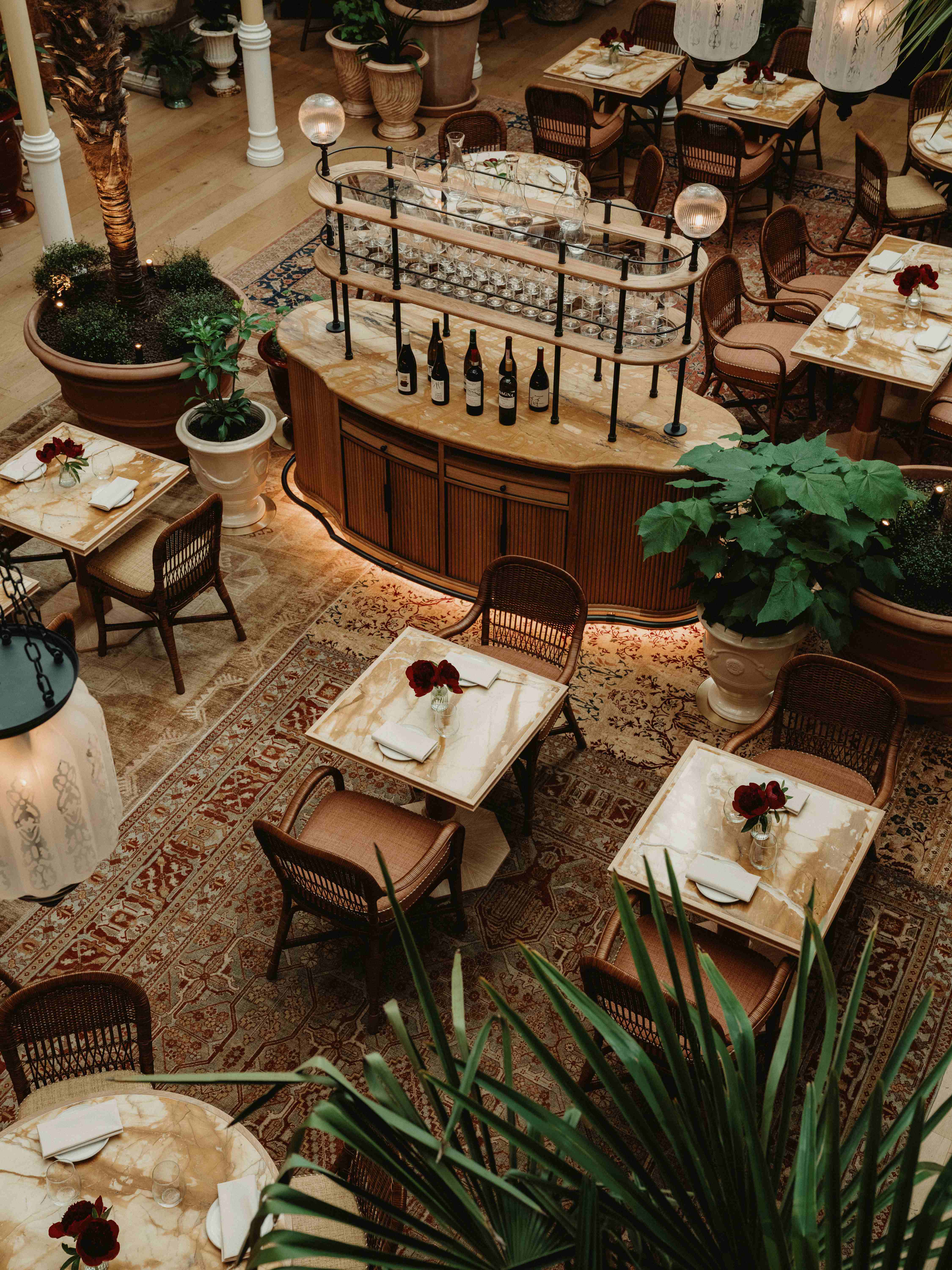 A dazzling restaurant with pendant glass and wrought iron chandeliers, wood and leather furnishings, marble table tops, red and cream patterned rugs, and retro-inspired furnishings, alongside thriving plants.
