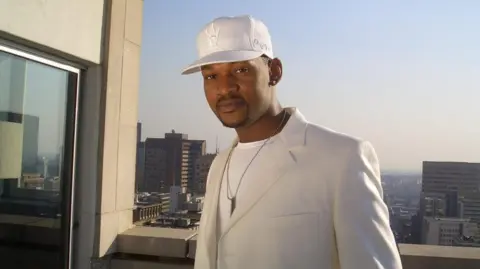 Shad Ellis A man stood on the roof of a building dressed in all-white. Cap, blazer and T-shirt. With a silver chain around his neck and a US city skyline behind him.