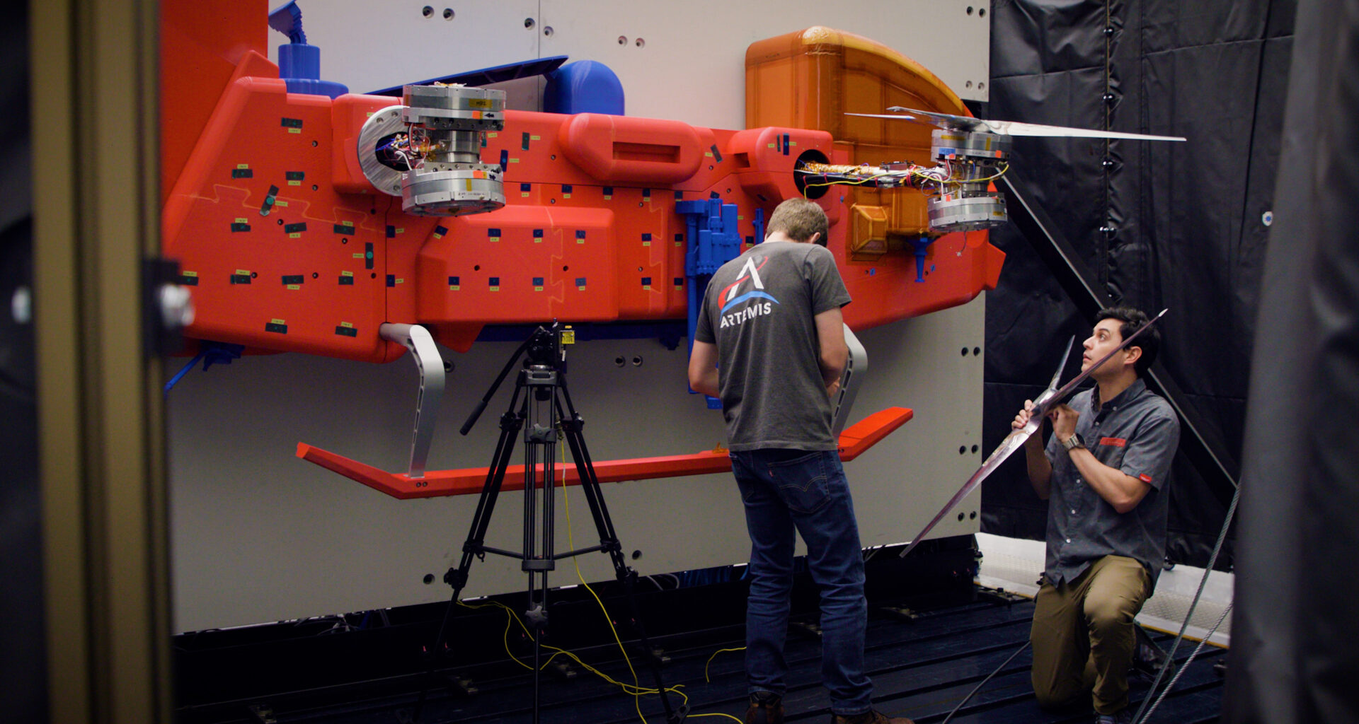 Two men in dark shirts work on a red car-sized rotorcraft protype in a testing chamber.