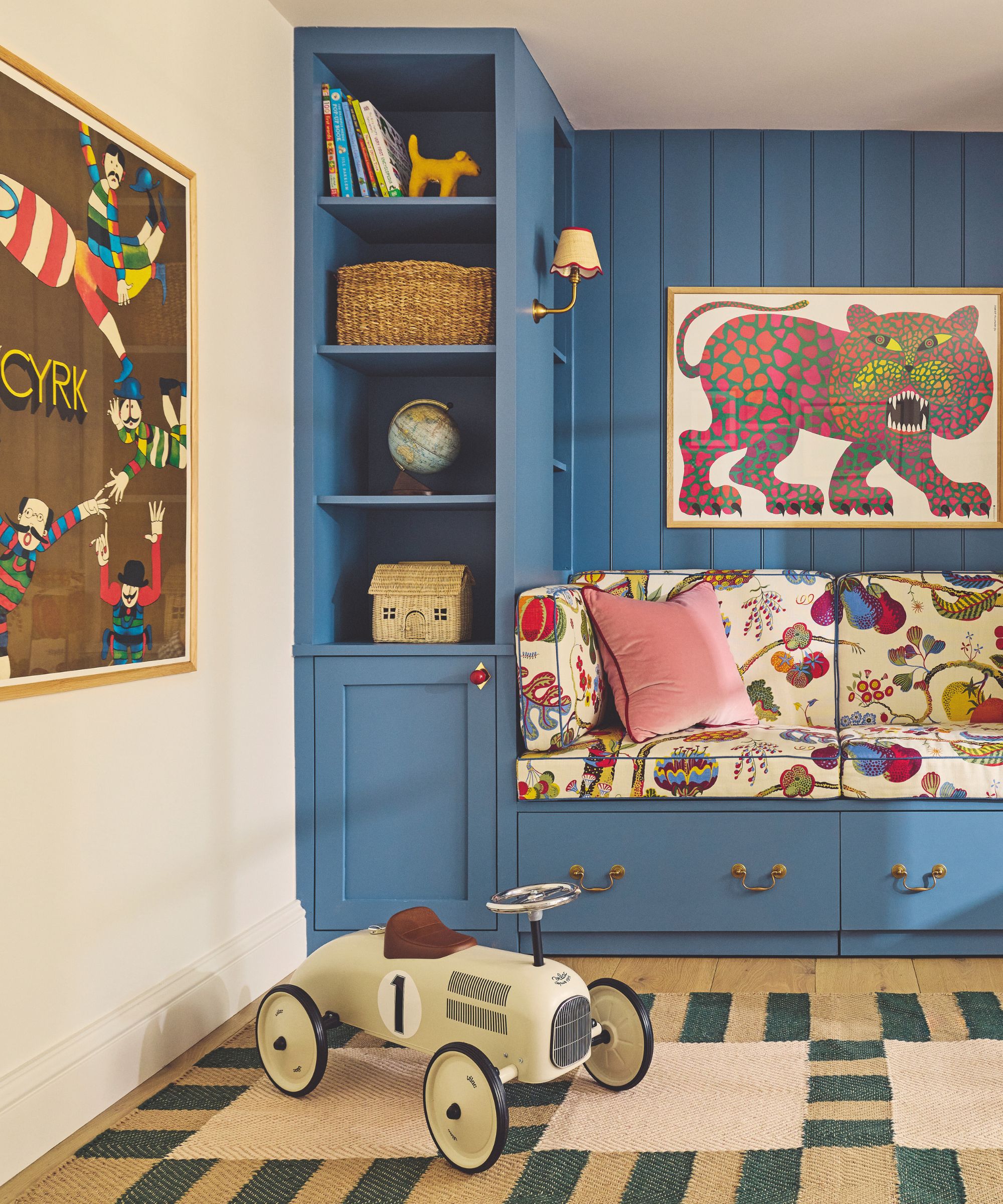 playroom with neutral walls, large green striped rug, and custom built in bench seat with shelves and cabinetry all painted in a bright blue and accessorized with vibrant fabric and art