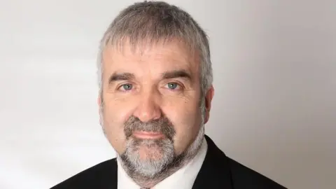 Handout A man looking at the camera, he has a grey/black beard and grey hair