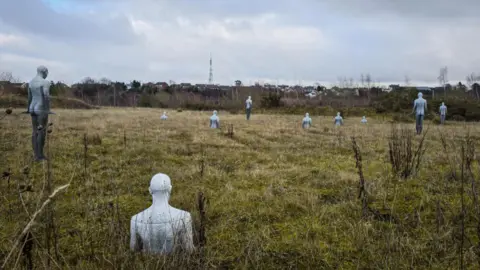 Lisa Rumsey A rural landscape with grassy fields and shrubs under a cloudy sky. Scattered across the field are several pale, human-shaped sculptures, some standing upright and others partially submerged in the ground.