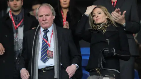 PA Media A man and woman stood side by side looking out onto a football game, with a look of concentration on both faces. The man is wearing a blue shirt and tie, whilst the woman is wearing a black coat and cheetah print scarf. 