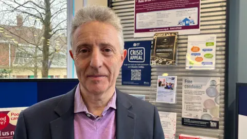 Nicola Haseler/BBC A man with grey hair stands in front of a health notice board in a pharmacy. The posters on the board promote local charities and health advice. 